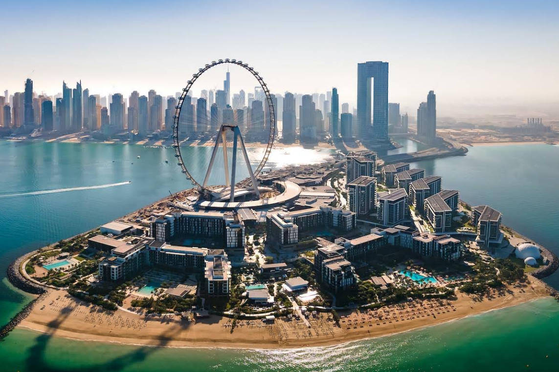 Aerial view of a modern island with a large Ferris wheel, luxury buildings, pools, and sandy beaches, with a city skyline and skyscrapers in the background across the water.