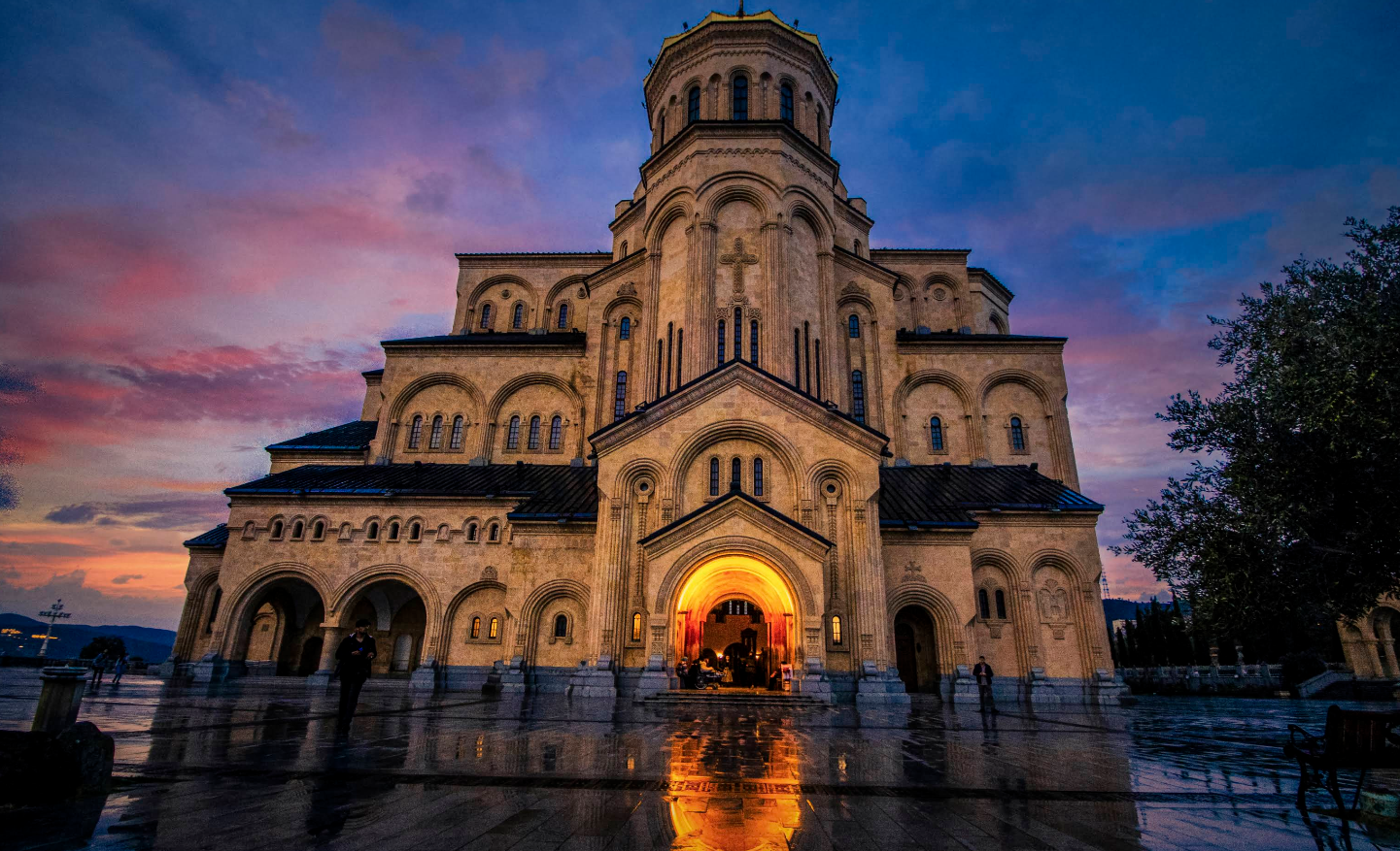 A large, illuminated cathedral with arched doorways stands against a colorful sunset sky, its reflection visible on wet pavement in front.
