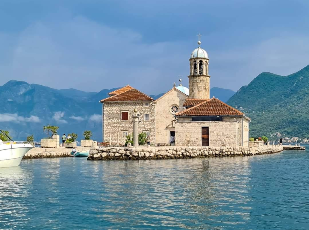 A stone church with a bell tower stands on a small island surrounded by calm water, with distant green mountains and a partly cloudy sky in the background.