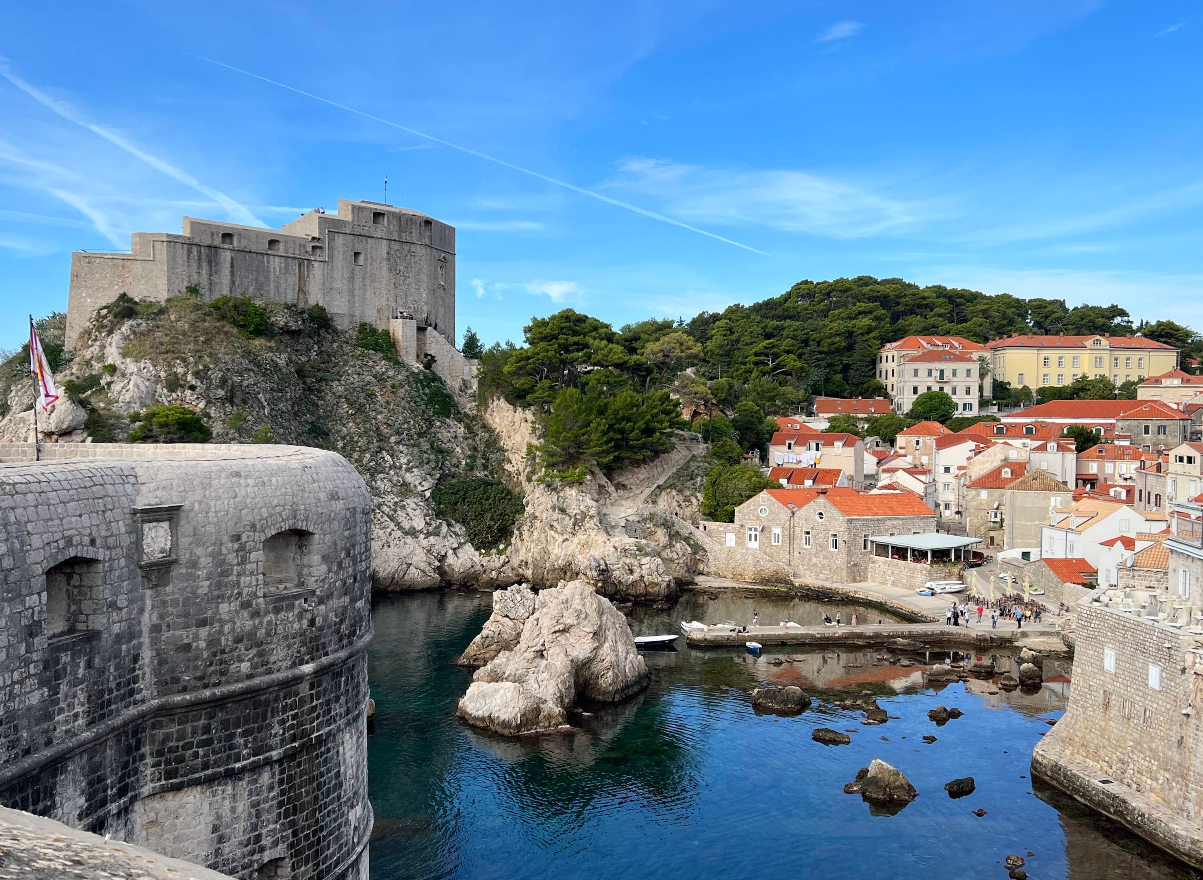 A stone fortress overlooks the blue sea, with rocky cliffs, red-roofed buildings, and green hills under a bright blue sky with wispy clouds.