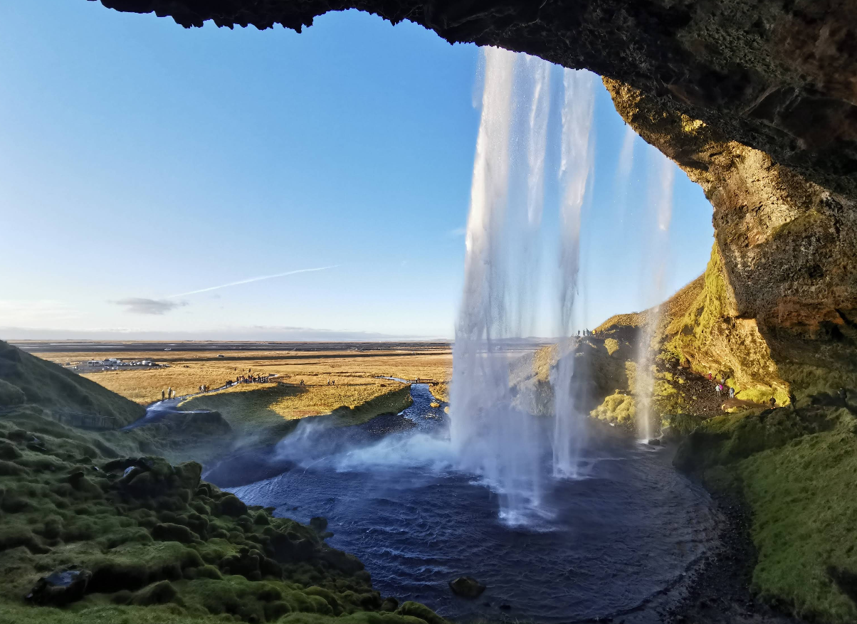 View from behind a tall waterfall cascading into a pool, with mossy rocks in the foreground and a bright, open landscape under a clear blue sky in the background.