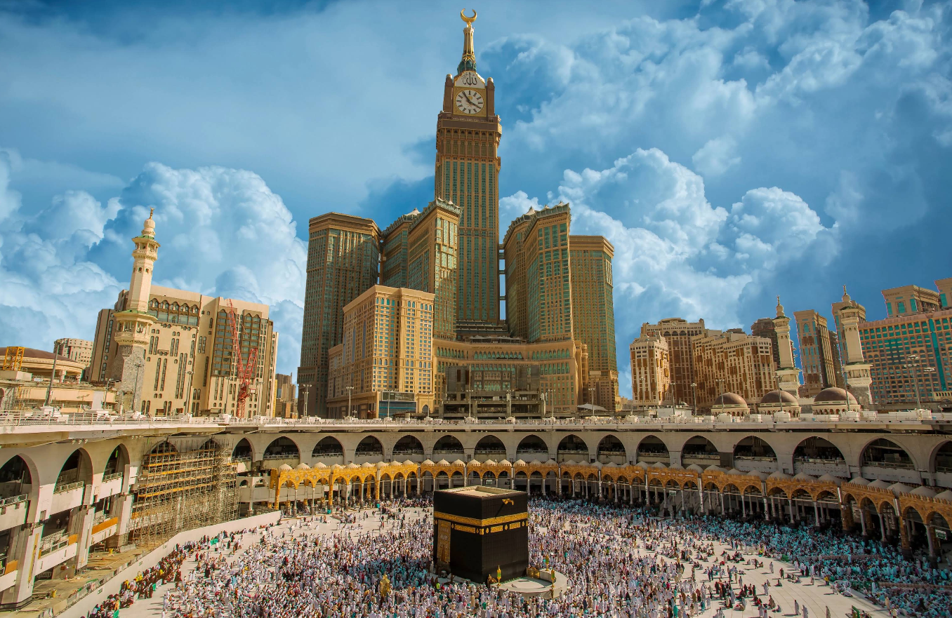 A large crowd gathers around the Kaaba at the Grand Mosque in Mecca, with the tall Abraj Al Bait clock tower and surrounding buildings visible under a dramatic, cloudy sky.
