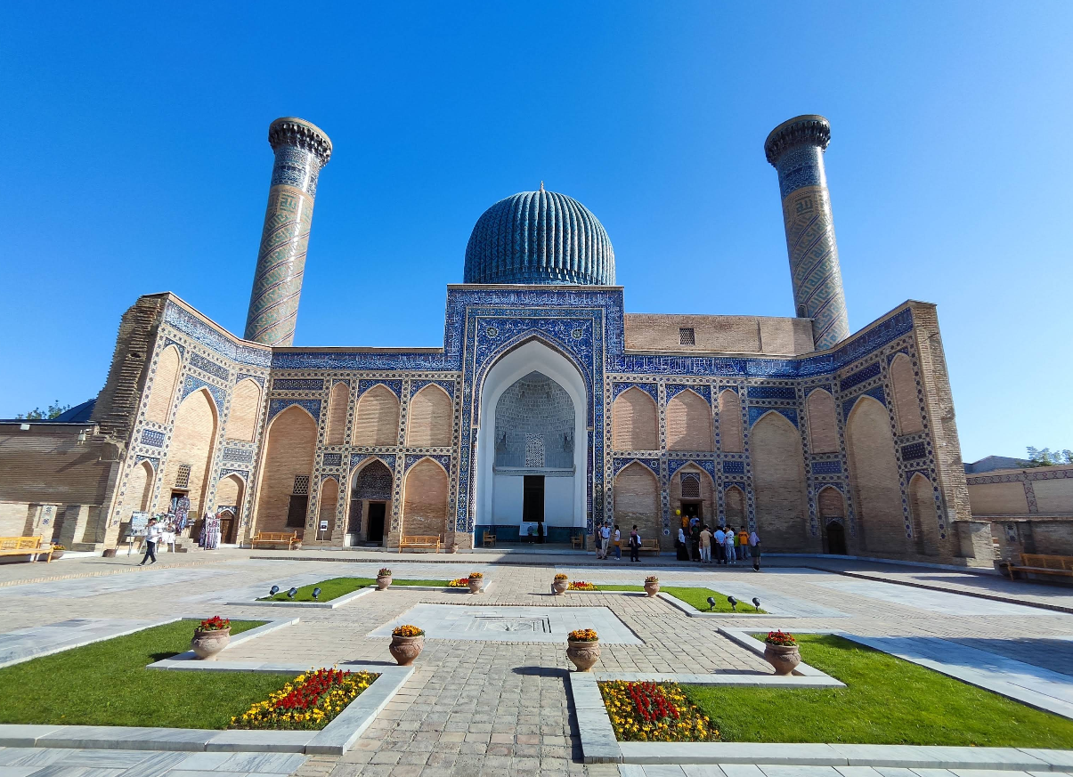 A grand mausoleum with a blue dome, arched entrance, tall minarets, and intricate tilework, surrounded by a courtyard with green lawns and flower beds under a clear blue sky.