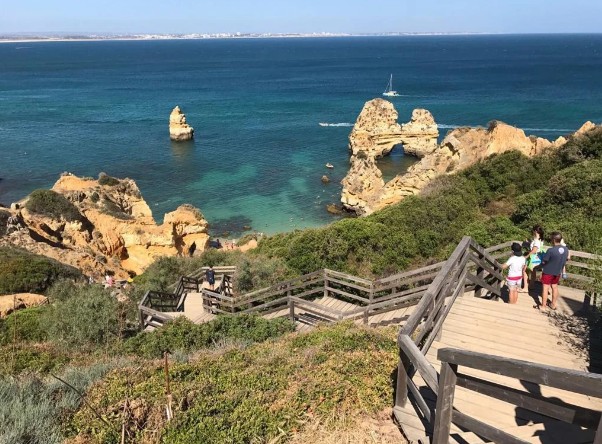 Wooden stairs lead down to a scenic coastline with rocky cliffs, clear blue-green water, and a sailboat in the distance. On this sunny day, several people are walking down toward the beach, ready to create travel experiences that went viral for their breathtaking beauty and unforgettable moments.