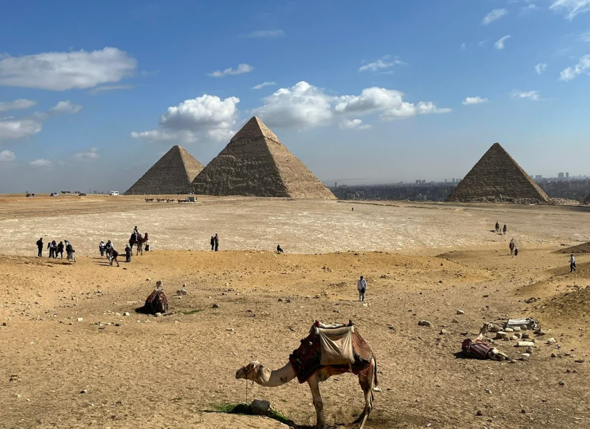 The Pyramids of Giza rise behind a sandy expanse. A camel rests in the foreground, while small groups of people walk across the desert, under a partly cloudy blue sky.