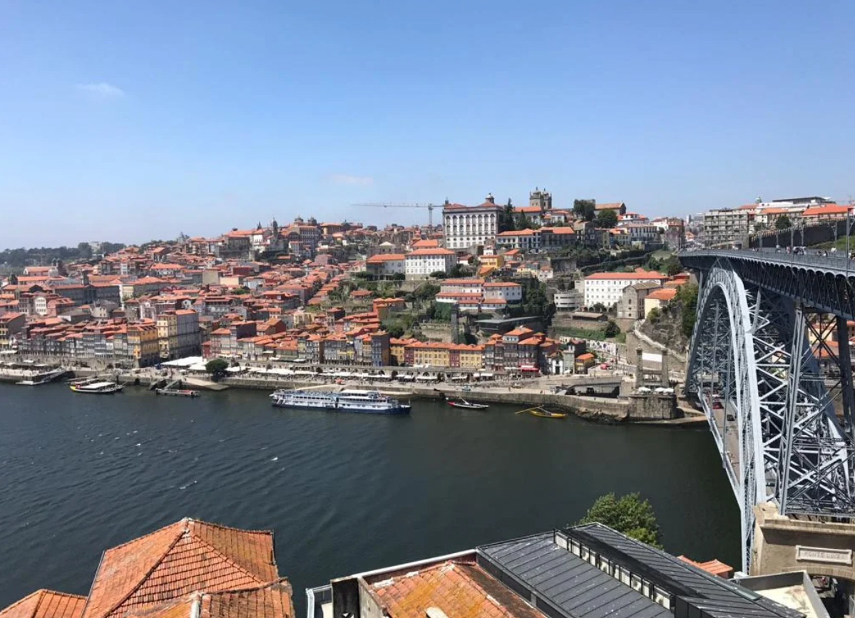 A cityscape of Porto, Portugal, shows red-roofed buildings on a hillside beside the Douro River, with the Dom Luís I Bridge on the right and boats docked along the waterfront—a picturesque setting often featured in travel experiences that went viral.