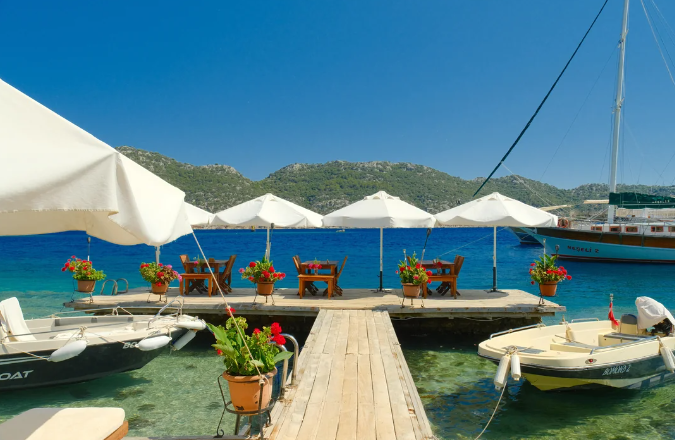 Wooden dock with tables and chairs under white umbrellas, surrounded by potted flowers, leading to blue sea with boats—a picturesque setting reminiscent of travel experiences that went viral. Green hills and clear sky provide the perfect backdrop.
