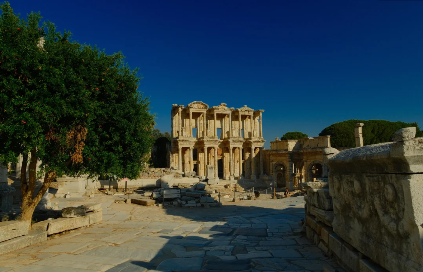 Ancient stone ruins with a large, ornate two-story structure in the center—surrounded by trees and clear blue sky—invite explorers to witness the awe-inspiring Library of Celsus in Ephesus, a destination renowned for travel experiences that went viral.