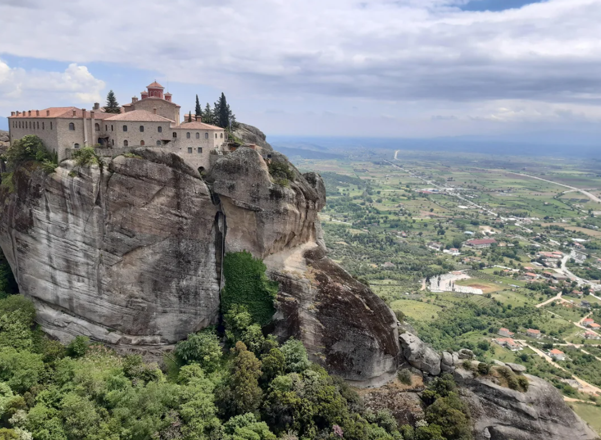 A monastery stands atop a tall, rocky cliff surrounded by lush greenery, overlooking a wide valley with fields and scattered buildings under a cloudy sky—an awe-inspiring scene reminiscent of travel experiences that went viral for their breathtaking beauty and sense of tranquility.