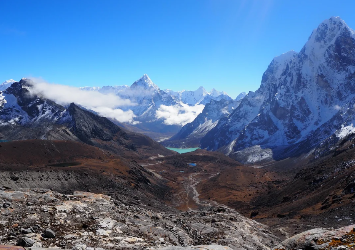 Snow-capped mountains under a clear blue sky, with a valley and a turquoise lake in the distance, surrounded by rocky terrain and sparse vegetation—scenery reminiscent of travel experiences that went viral for their breathtaking beauty.