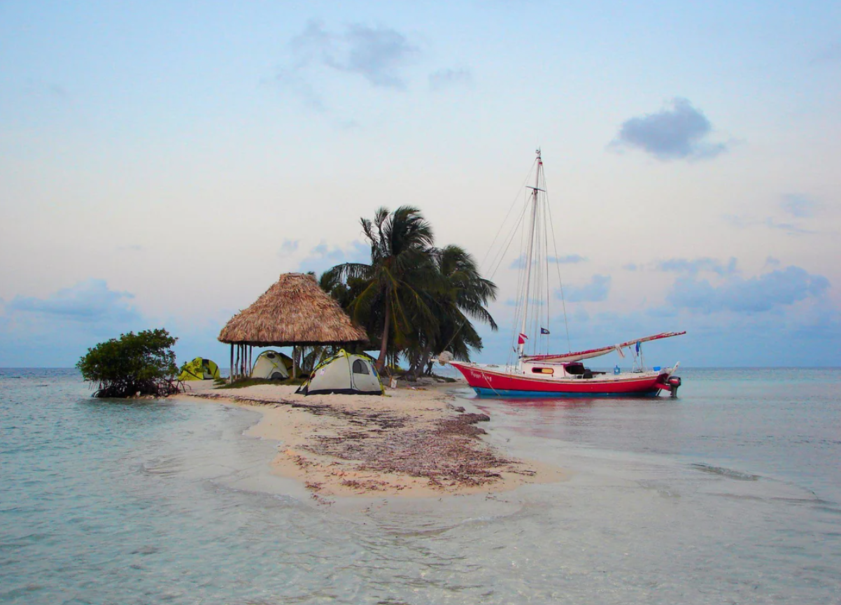 A small tropical island with palm trees, two tents, a thatched hut, and a red-and-white sailboat anchored nearby on calm, blue water at sunset—just the kind of paradise featured in travel experiences that went viral.