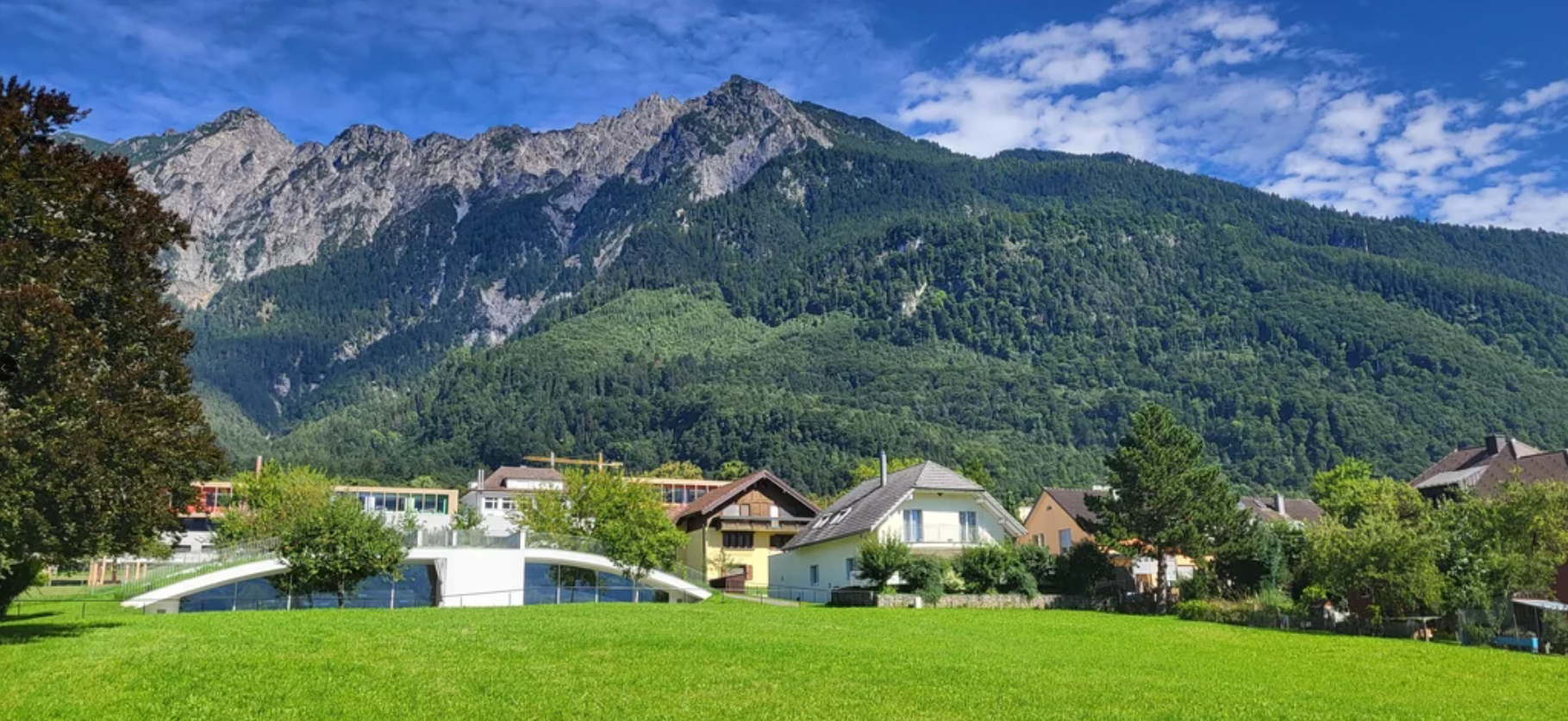 A green field with houses, trees, and a small white bridge in front of a forested mountain range under a blue sky with scattered clouds—an idyllic landscape reminiscent of travel experiences that went viral for their breathtaking scenery and serene beauty.