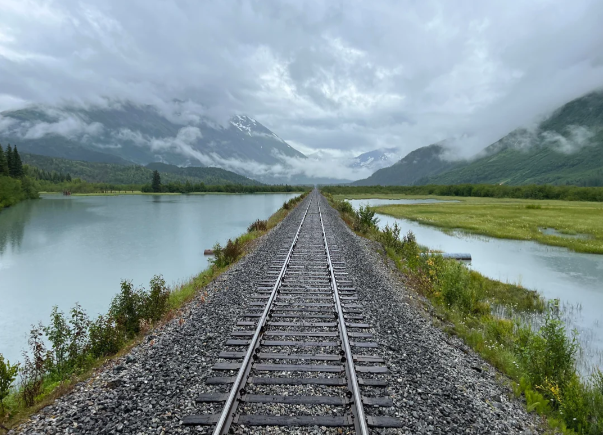 Railroad tracks stretch into the distance through lush greenery and over water, with mountains and cloudy skies in the background.