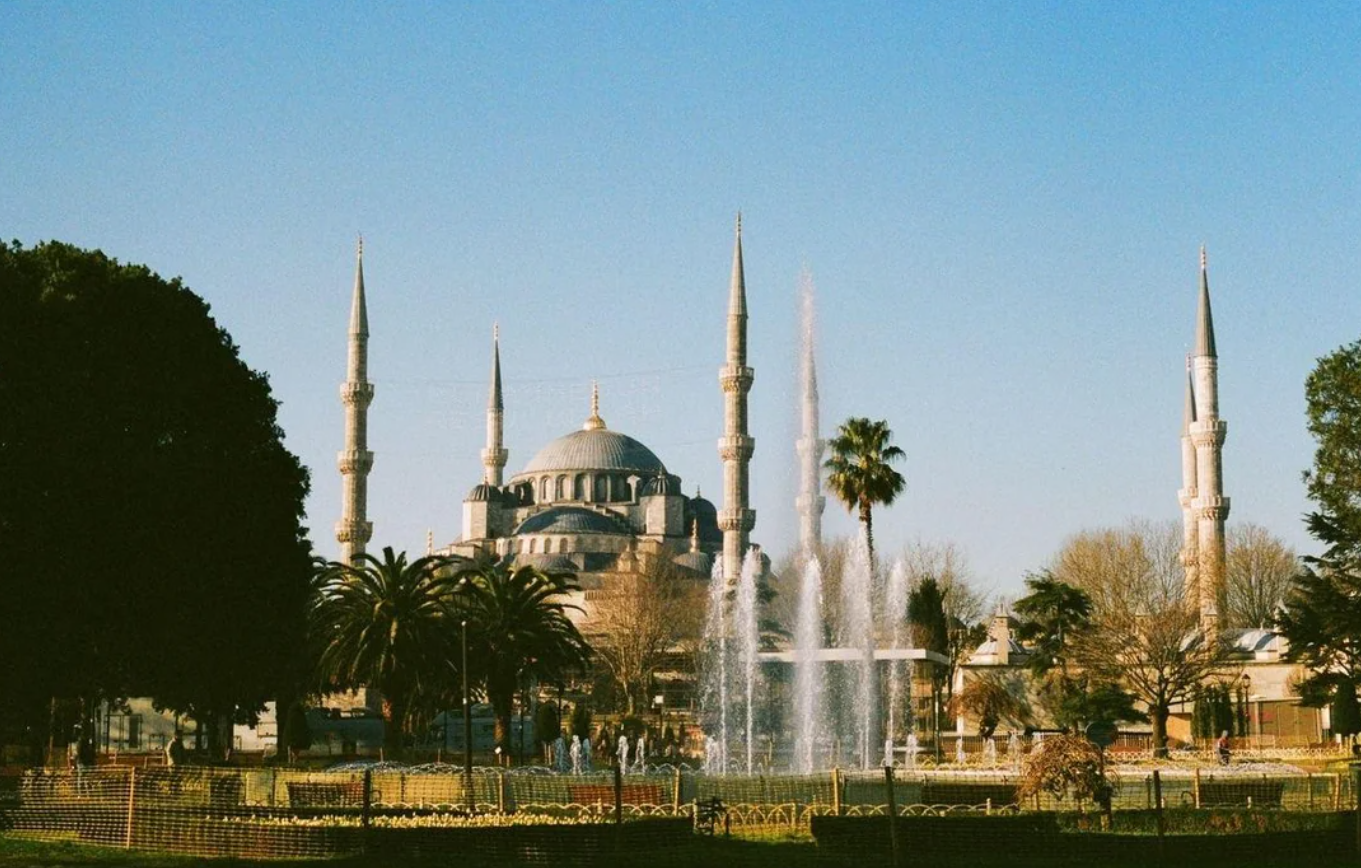 The Blue Mosque in Istanbul stands with six minarets under a clear blue sky, surrounded by trees and fountains in the foreground.