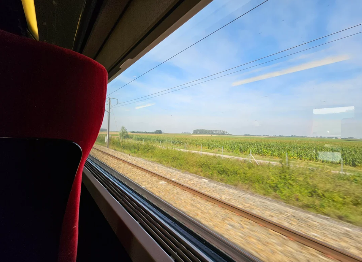 View from a train window showing railway tracks, grassy fields, and a blue sky; part of a red train seat is visible in the foreground on the left.