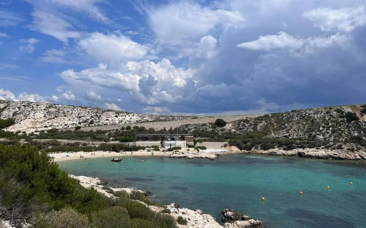 A small beach with turquoise water, rocky hills, and green shrubs. A few people are on the sandy shore near a building. Bright clouds fill the blue sky above the scenic coastline.