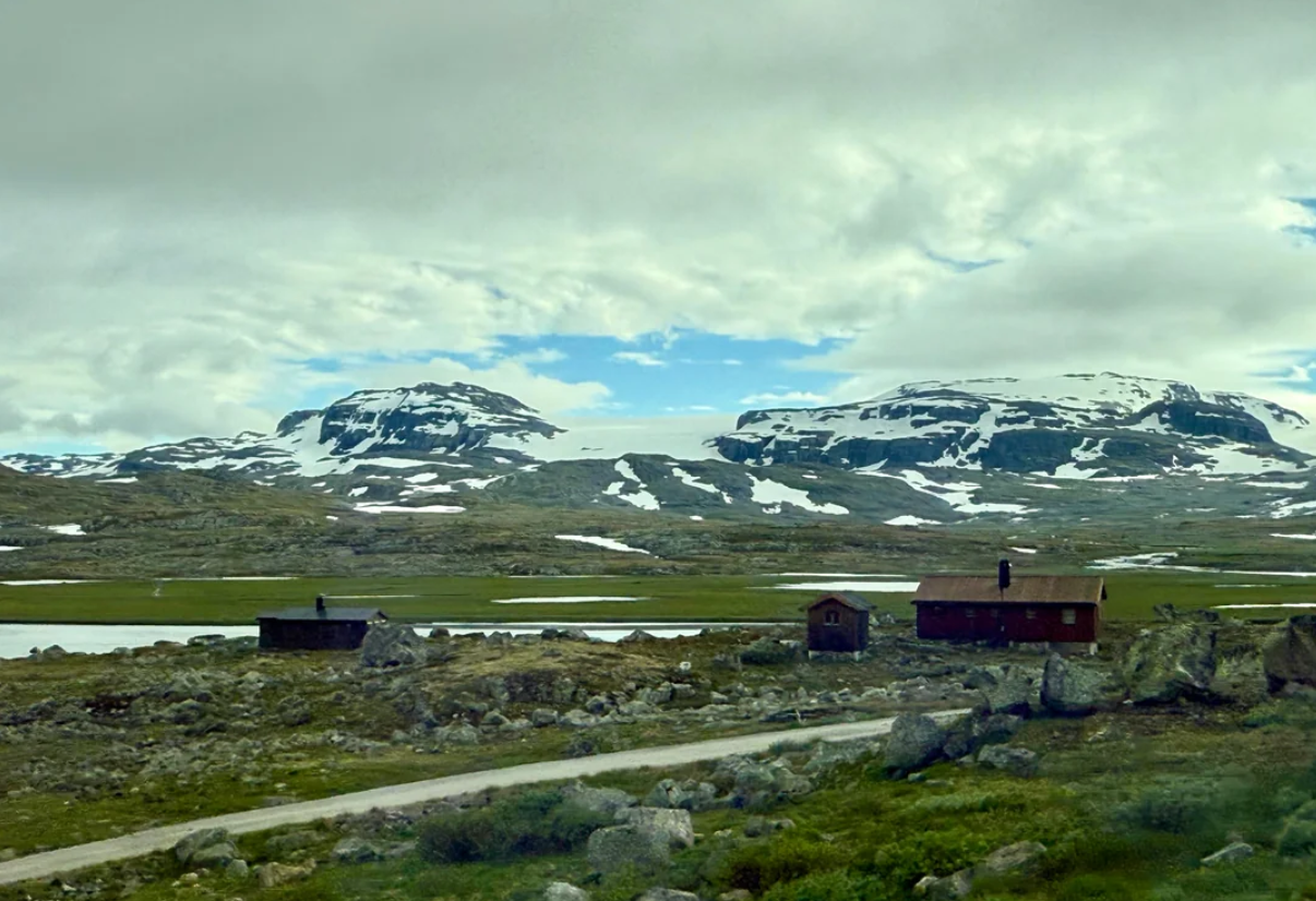 Wooden cabins sit on rocky terrain with patches of grass, in front of snow-capped mountains under a cloudy sky. A gravel road runs through the foreground.