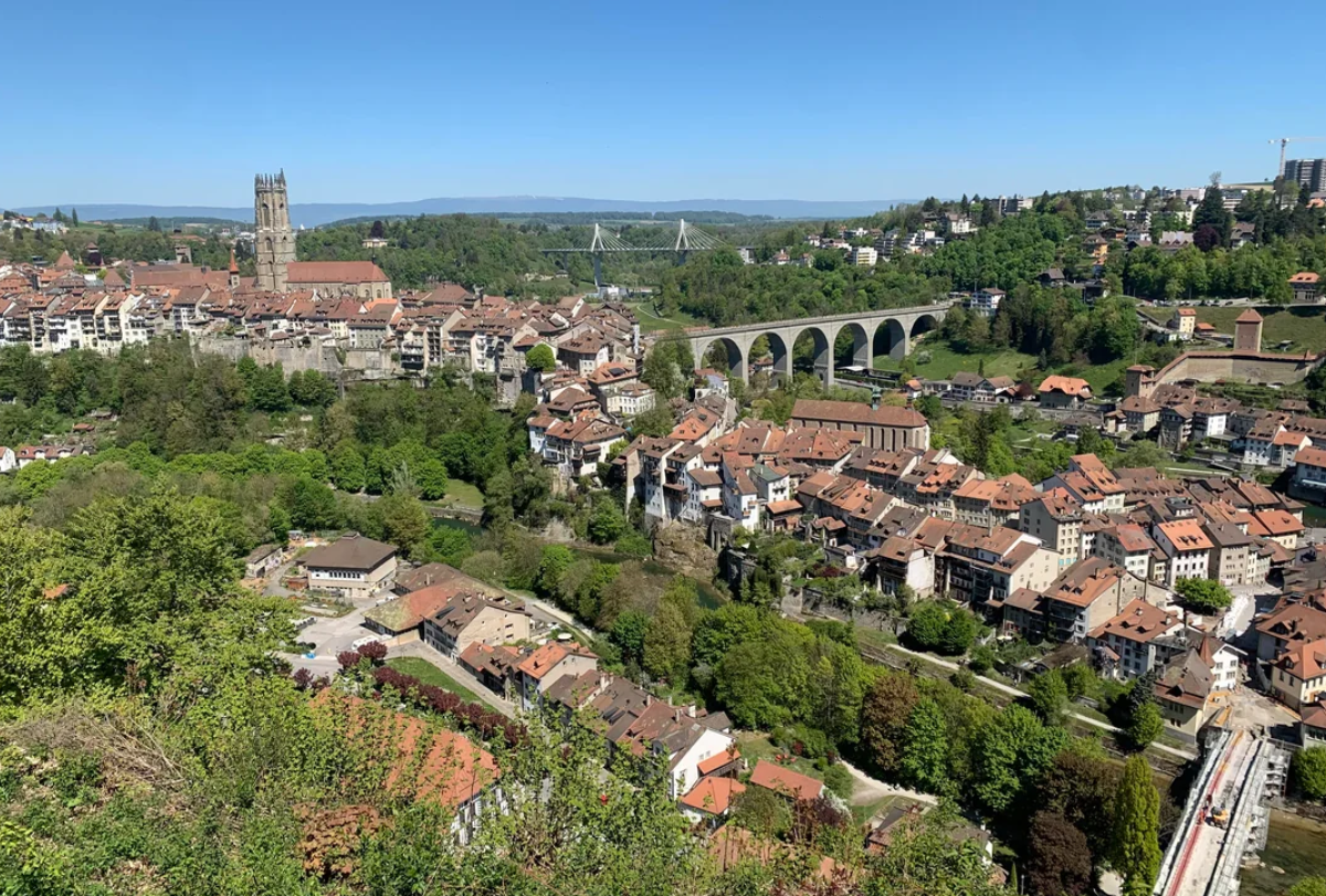 A scenic view of a European town with red-roofed buildings, a large stone viaduct, a tall cathedral, and lush green trees under a clear blue sky.