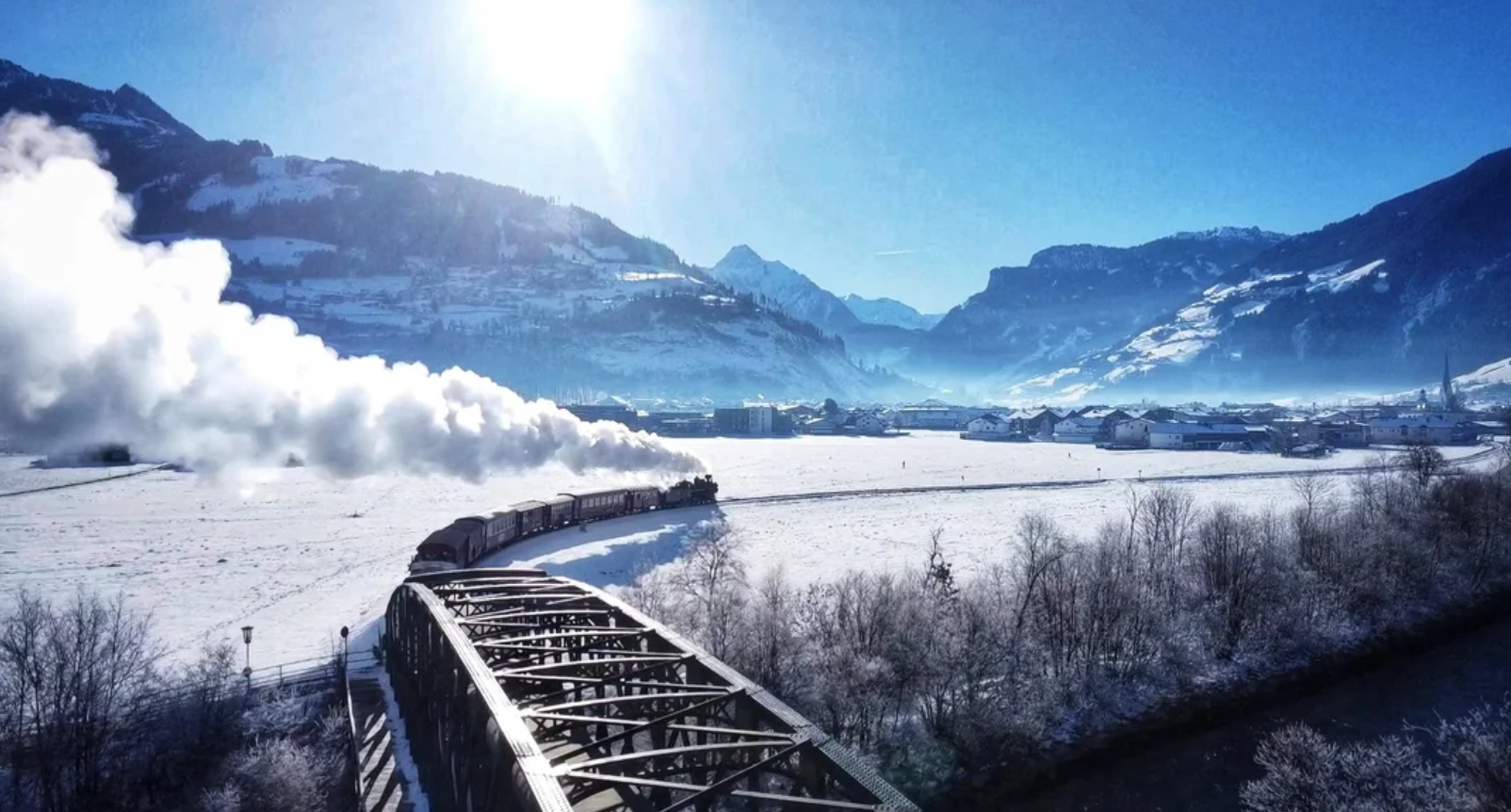 A steam train crosses a snowy landscape on a metal bridge, smoke trailing behind. Snow-covered mountains and a bright sun are visible under a clear blue sky.