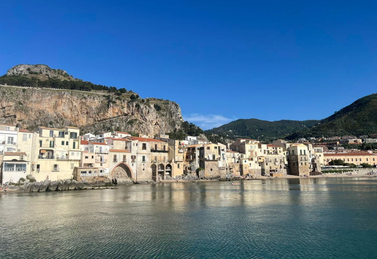 Colorful buildings line the waterfront beneath rocky hills in a coastal town, reflecting on calm water under a clear blue sky.
