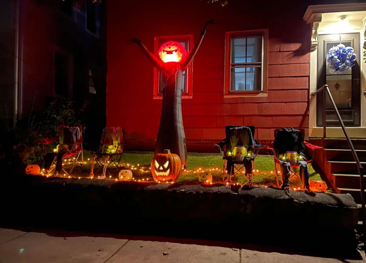 A yard decorated with glowing pumpkins, string lights, and a tall inflatable figure with a jack-o’-lantern head and outstretched arms showcases festive Halloween yard decorations. Four chairs are set up near the decorations in front of a red house at night.