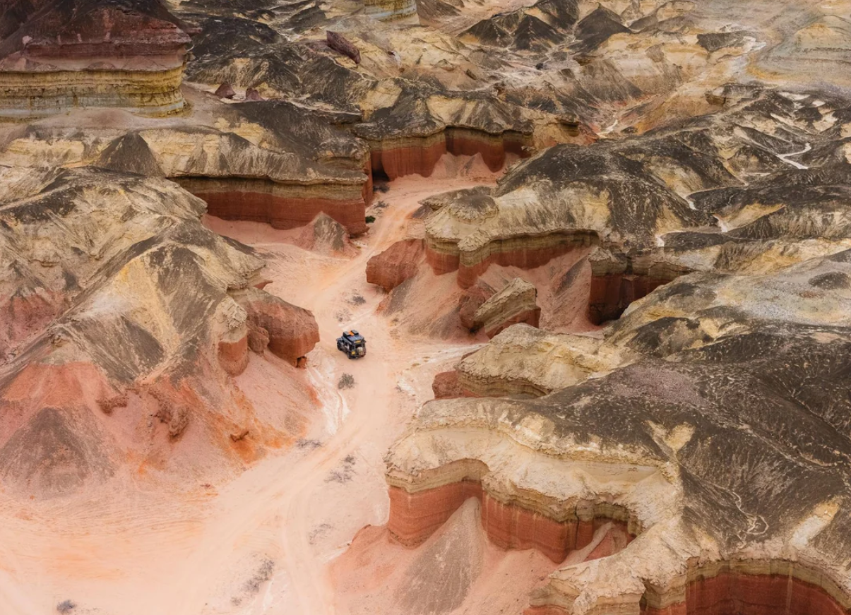 An aerial view of a rugged, multicolored canyon landscape with layered rock formations and a vehicle driving along a sandy road through the canyon.