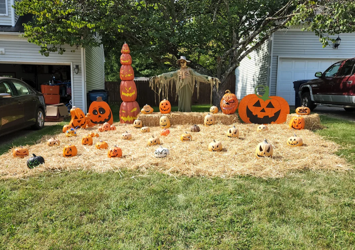 A yard decorated for Halloween with many carved pumpkins, a tall stacked pumpkin display, a scarecrow, and hay bales in front of a house and cars showcases festive Halloween yard decorations.