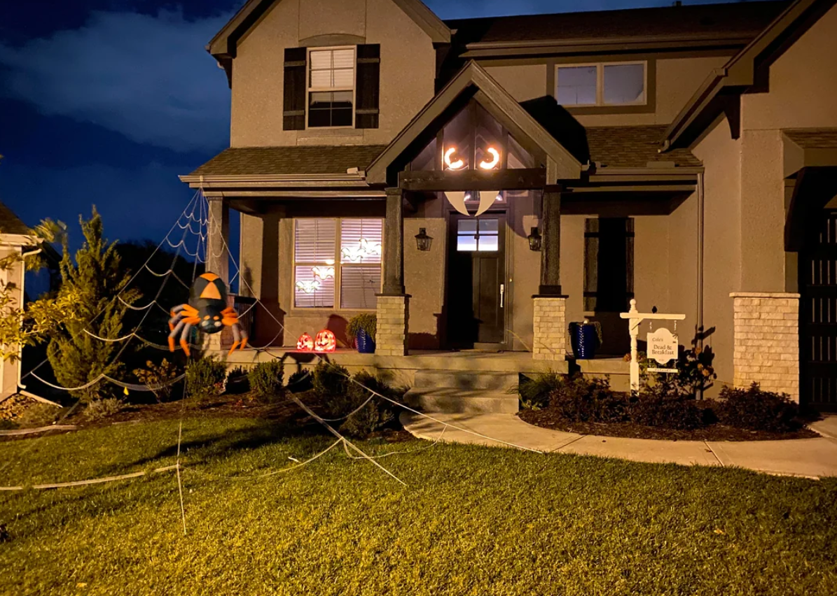 A house decorated for Halloween at night with glowing eyes in the window, a giant spider and web on the lawn, pumpkins on the porch, and orange string lights inside creates a perfect showcase of Halloween yard decorations.