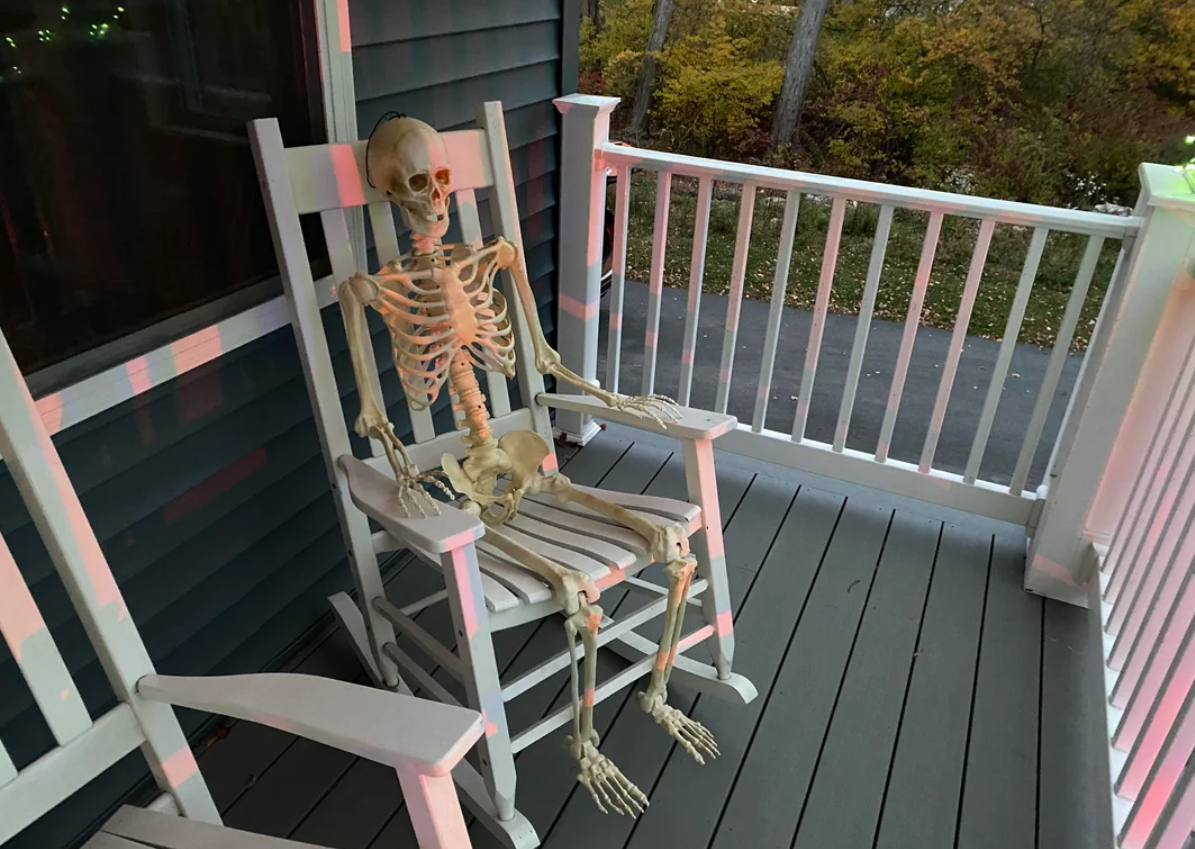 A plastic skeleton sits upright in a white rocking chair on a porch with gray flooring and white railings, making it a perfect addition to your Halloween yard decorations. Trees with autumn leaves are visible in the background.