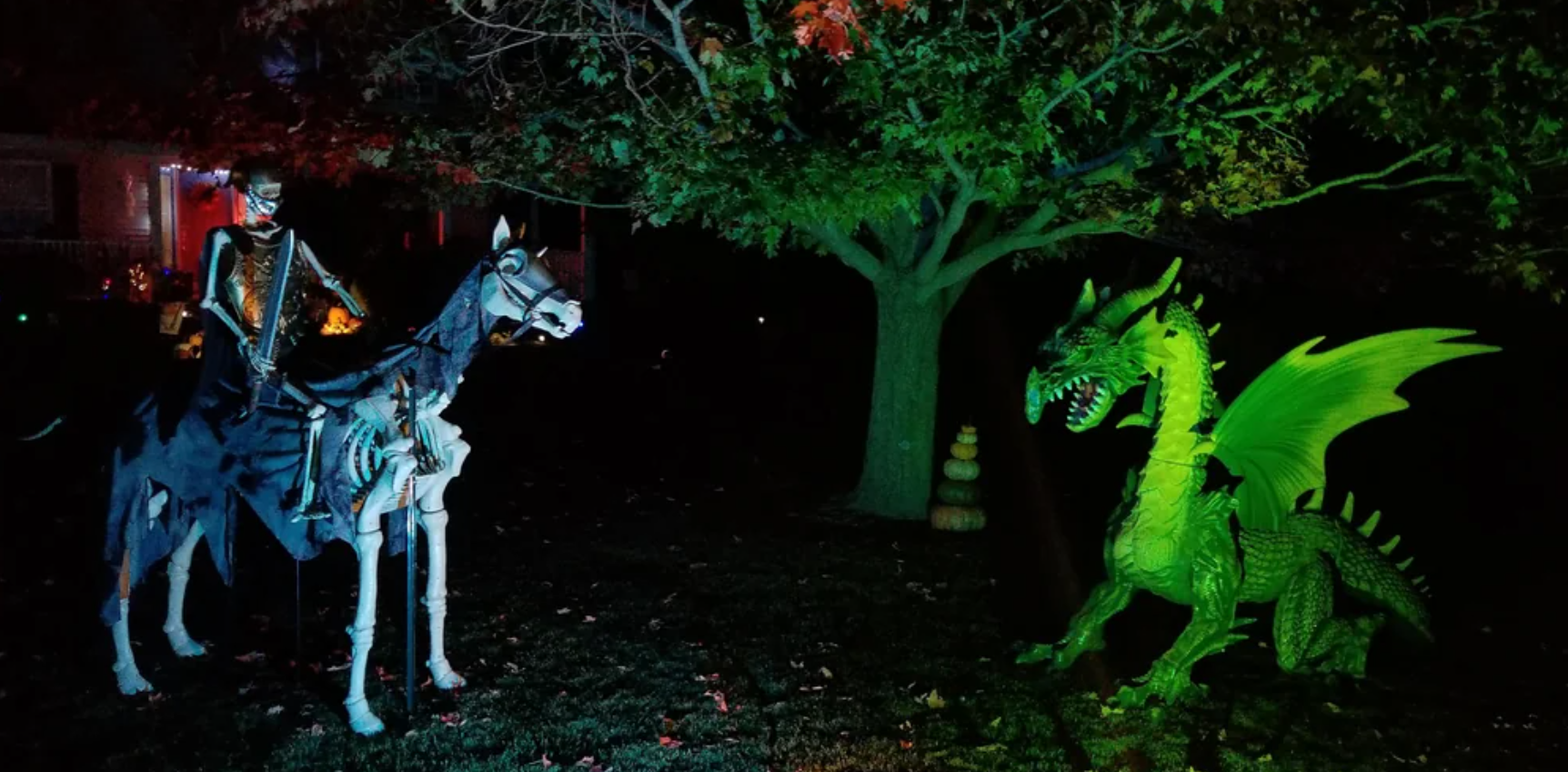 A skeleton figure on a skeletal horse faces a large green dragon under tree branches at night, both illuminated with colored lights for a spooky scene—perfect for standout Halloween yard decorations.