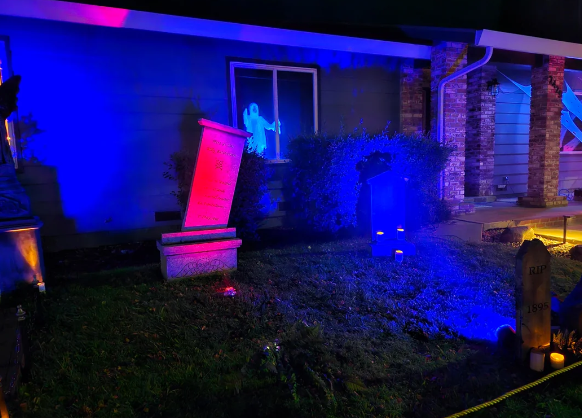 A house yard at night with tombstones, colored lights, and a ghostly figure projected in a window, creating a spooky Halloween scene enhanced by eye-catching Halloween yard decorations.