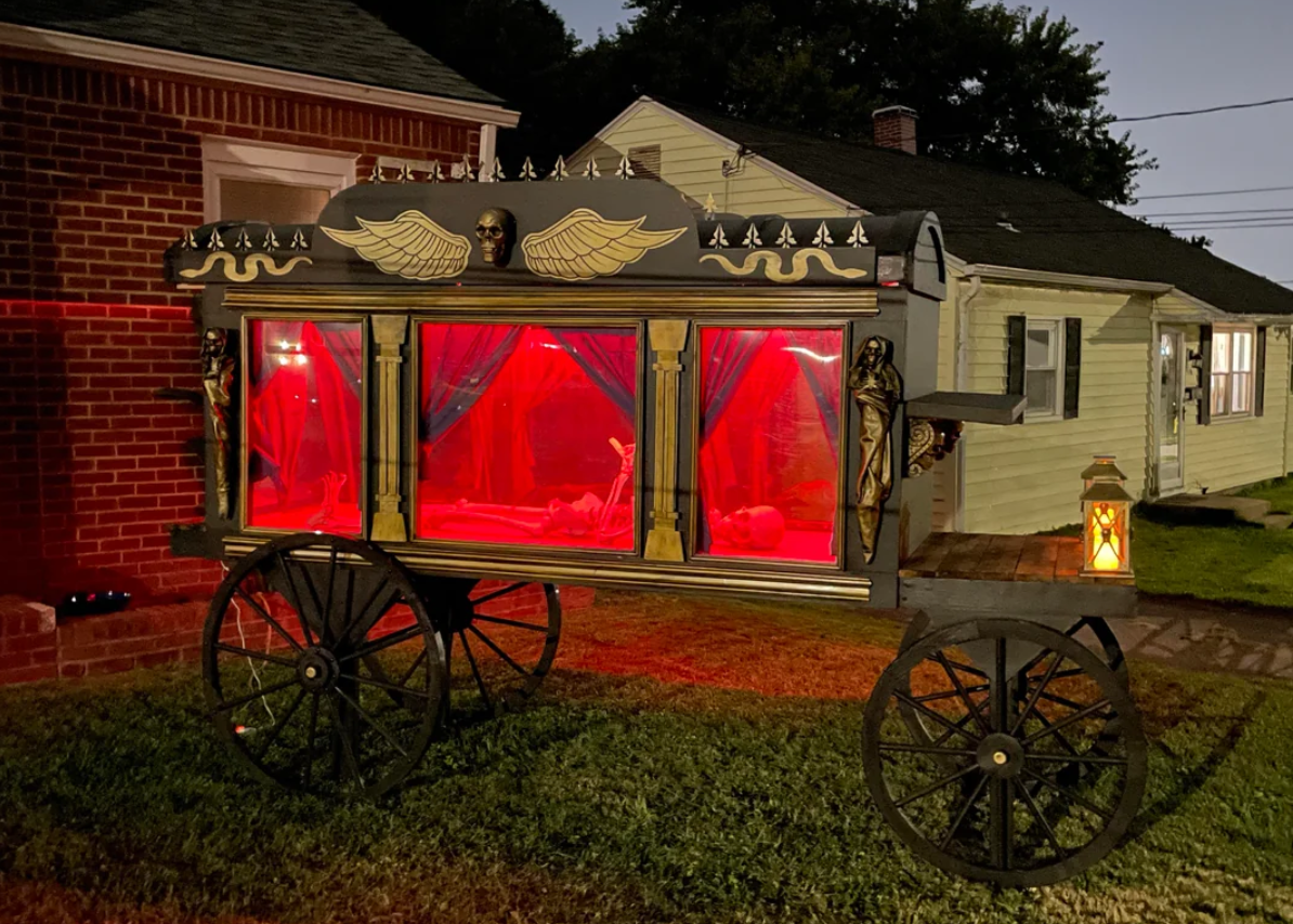 A vintage black and gold funeral hearse wagon with red-lit windows and Egyptian-style decorations, perfect as Halloween yard decorations, sits on grass near brick and white houses at dusk.