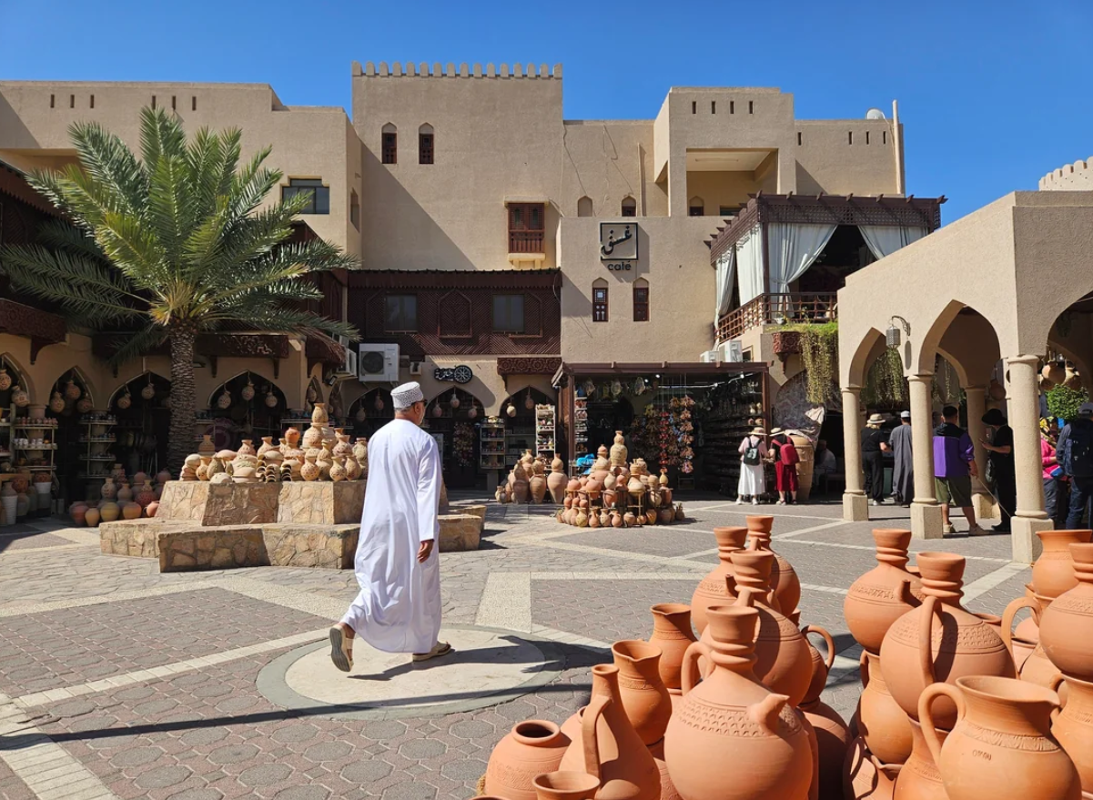 A person in traditional white attire walks through a sunny outdoor market with many clay pots, palm trees, and beige buildings with arches and balconies in the background.