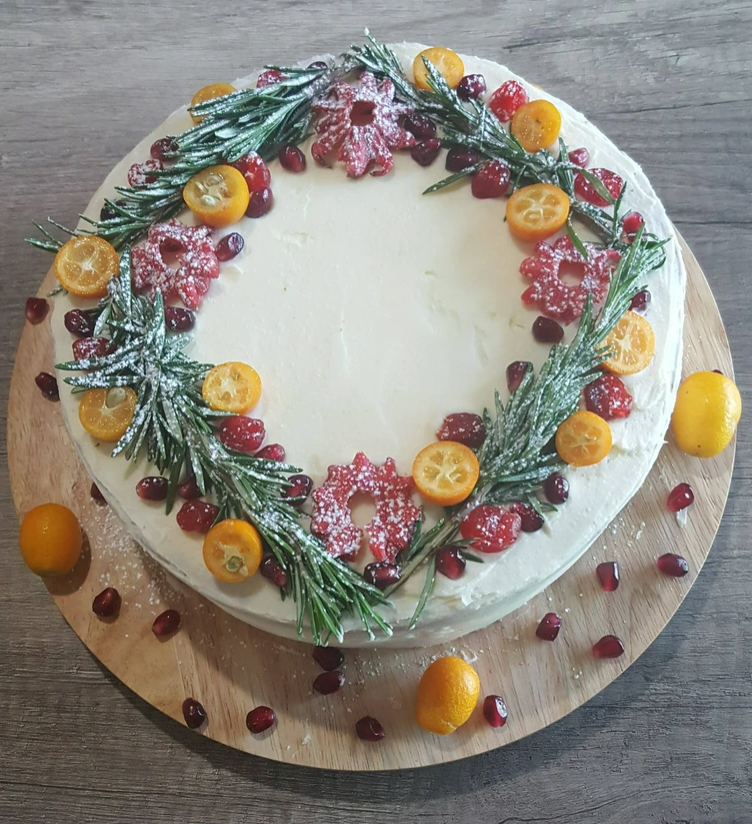 Round cake with white frosting, topped with kumquats, cranberries, sugared rosemary, pomegranate seeds, and snowflake-shaped cookies arranged in a wreath pattern—a delightful centerpiece for your homemade Christmas treats. Decorated with festive fruits on a wooden board.