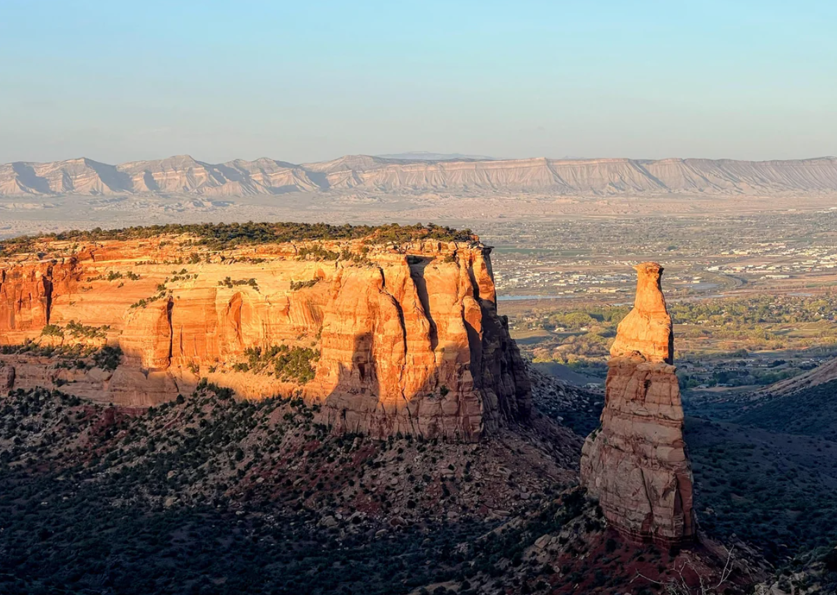 Sunlit red rock cliffs and a tall spire rise above a green valley, with distant layered mountains under a clear blue sky in the background.