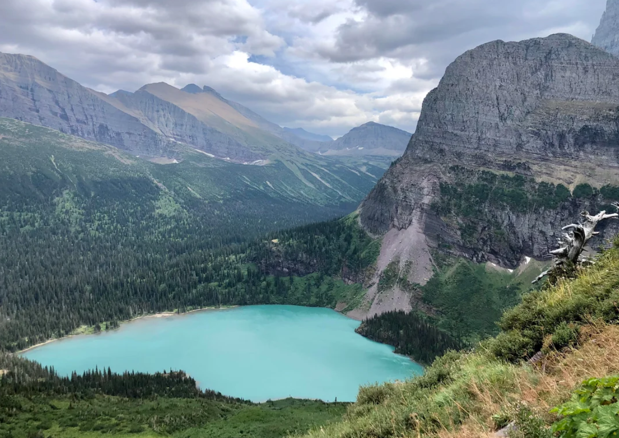 A turquoise lake surrounded by dense green forest and mountains under a cloudy sky, with rocky cliffs rising on the right side of the image.