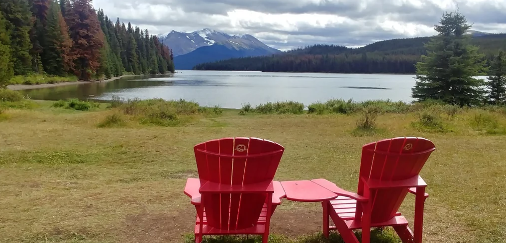 Two red Adirondack chairs face a calm lake, surrounded by forest with mountains in the background under a cloudy sky.