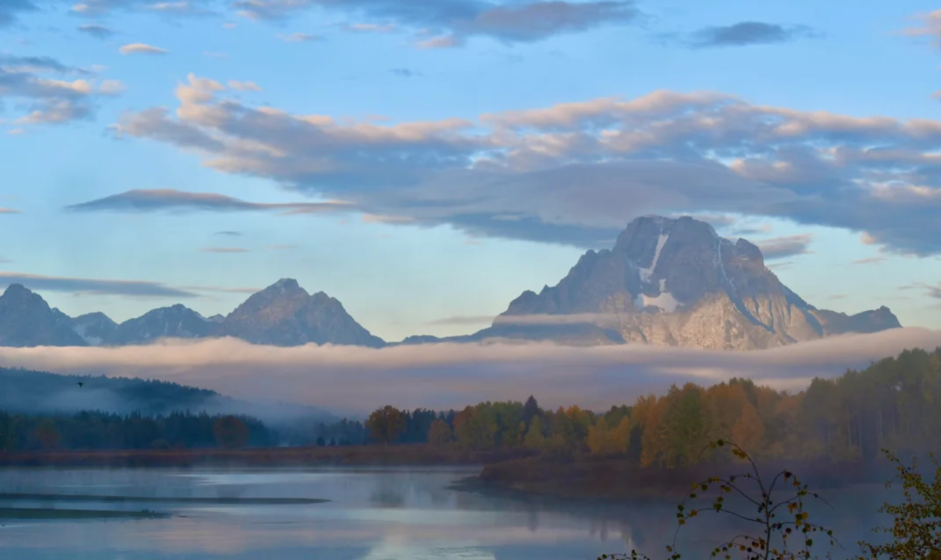 Snow-capped mountains rise above a misty lake, bordered by autumn trees. The sky is partly cloudy, reflecting soft blue and orange hues on the water.