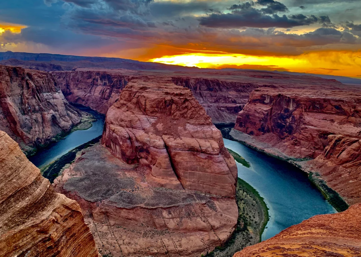 A dramatic sunset over Horseshoe Bend, where the Colorado River curves around a massive red rock formation surrounded by steep canyon walls.