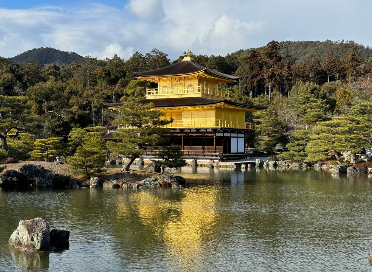 A golden pavilion sits by a tranquil pond surrounded by trees and mountains, reflecting on the water under a partly cloudy sky.