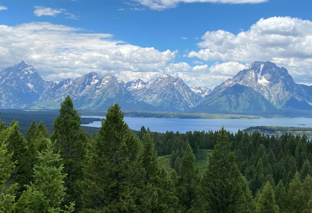 Snow-capped mountains under a partly cloudy sky, with a lake and dense evergreen forest in the foreground—this breathtaking scene captures the natural beauty often discovered on trips to national parks in America.