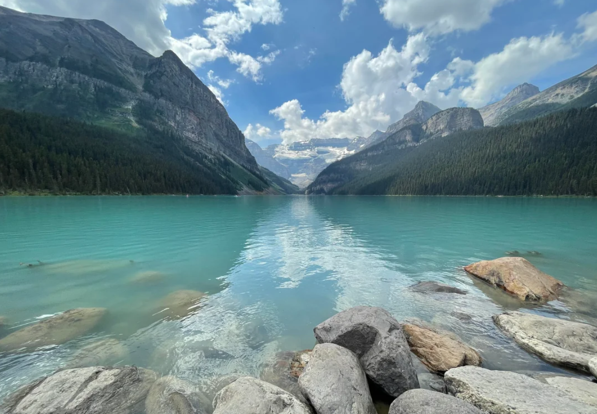 Turquoise lake surrounded by rocky shoreline, dense evergreen forests, and tall mountains under a partly cloudy blue sky—perfect scenery for unforgettable trips to national parks in America.