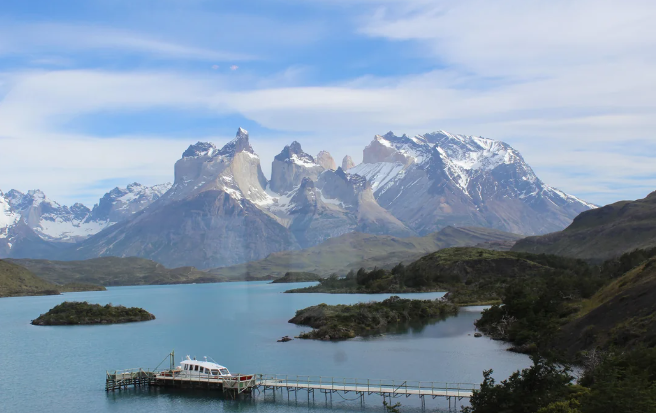 A boat docked at a pier on a calm lake, surrounded by green hills and snow-capped mountains in the background beneath a partly cloudy sky—an inviting scene reminiscent of trips to national parks in America.