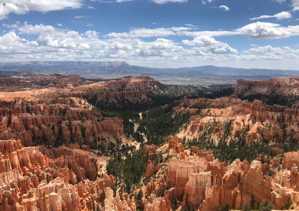 View of Bryce Canyon with orange rock formations, scattered green pine trees, and a partly cloudy blue sky stretching over distant mountains—an iconic landscape perfect for unforgettable trips to national parks in America.