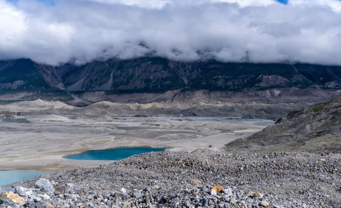 Rocky landscape with scattered stones in the foreground, a small blue lake in the midground, and misty mountains partially covered by clouds in the background, reminiscent of the dramatic vistas found in American National Parks.