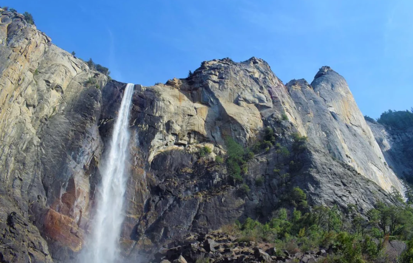 A tall waterfall cascades down a rugged, rocky cliffside surrounded by sparse green trees under a clear blue sky, reminiscent of the breathtaking landscapes found in American National Parks.