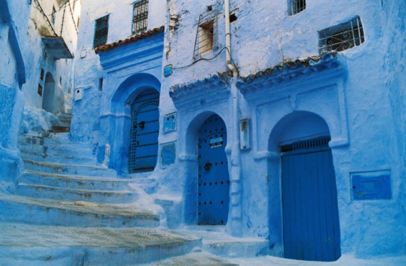 A narrow stone staircase leads past blue-painted doors and arches on buildings in Chefchaouen, Morocco, known as the Blue City. The scene features vibrant blue walls and intricate architectural details.