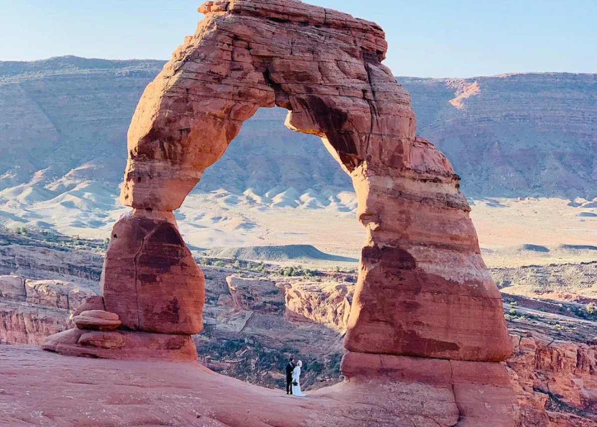 A couple stands beneath Delicate Arch, one of the most iconic features of American National Parks—a large, freestanding red rock arch in Arches National Park, Utah, with mesas and desert landscape in the background.