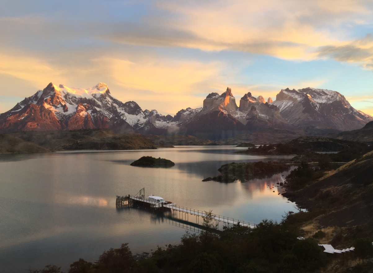 Snowy mountains reflect in a calm lake at sunset, with a small dock and building on the water, set amidst rugged terrain and soft clouds in the sky—a serene scene reminiscent of the breathtaking landscapes found in American National Parks.
