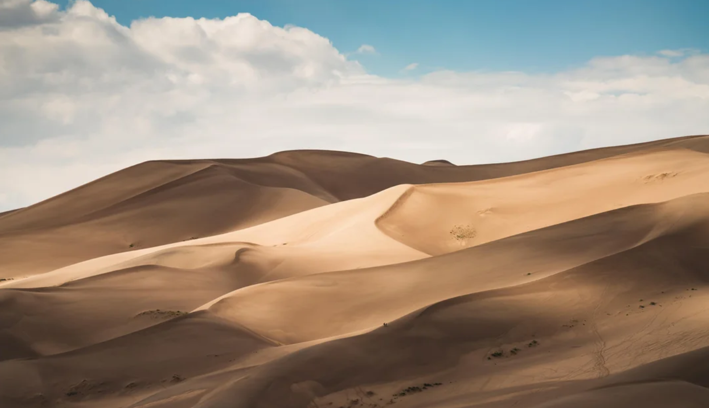 Rolling sand dunes under a partly cloudy sky, with sunlight casting highlights and shadows on the smooth desert landscape—a scene reminiscent of the breathtaking vistas found in American National Parks.